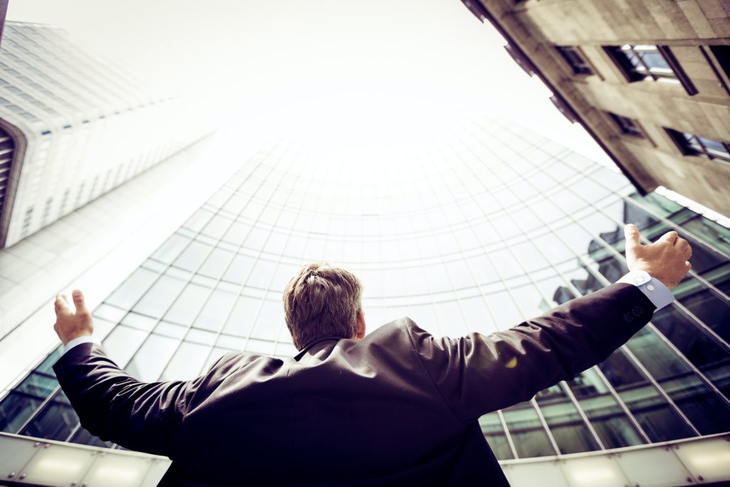 Man standing next to skyscraper with his arms aimed at the sky as if he has succeeded