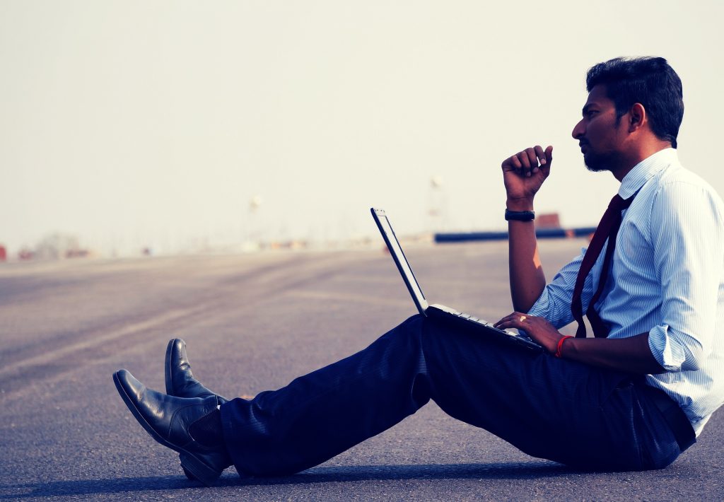 A man sitting on the pavement wearing a shirt and writing on his computer adapting to his surroundings