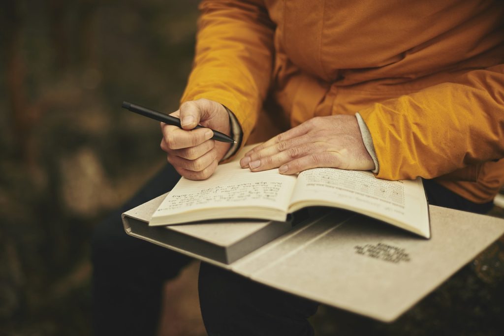 A person in a yellow shirt with two books in it's lap journaling in one of the books using self-reflection journal prompts